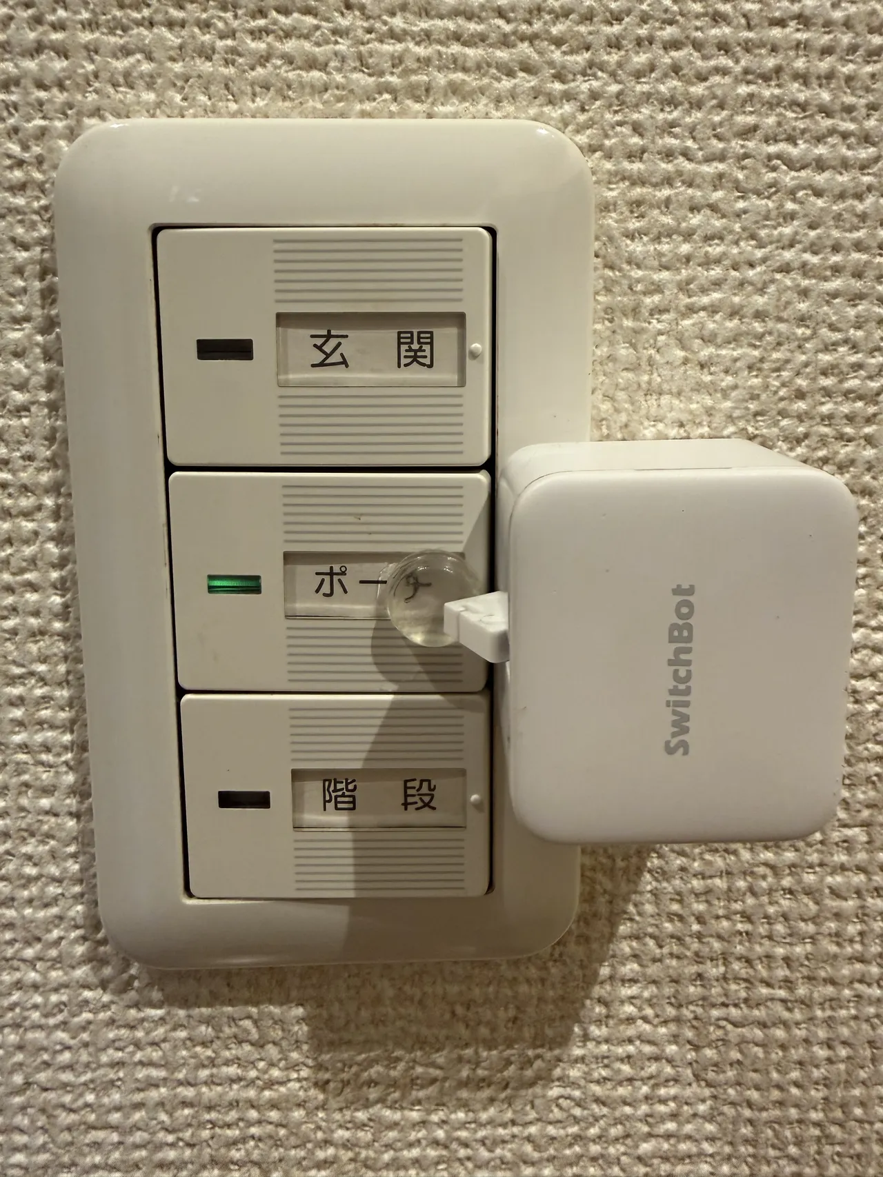 壁のスイッチプレート。「玄関」「ポーチ」「階段」の 3 つのスイッチが縦に並び、白い SwitchBot ボットがプレートの右側に貼られ、アームを「ポーチ」のスイッチに向けている。「ポーチ」の表示灯は緑