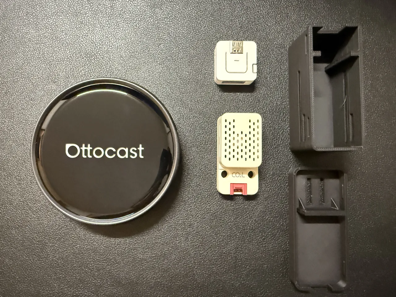 黒い革の上に並べた部品。左に丸い Ottocast の Android ボックス、中央の上に白い ATOMS3 Lite、その下に穴の並んだ SCD41 ユニット、右に 3D プリンタで作った黒いケースの本体とふた
