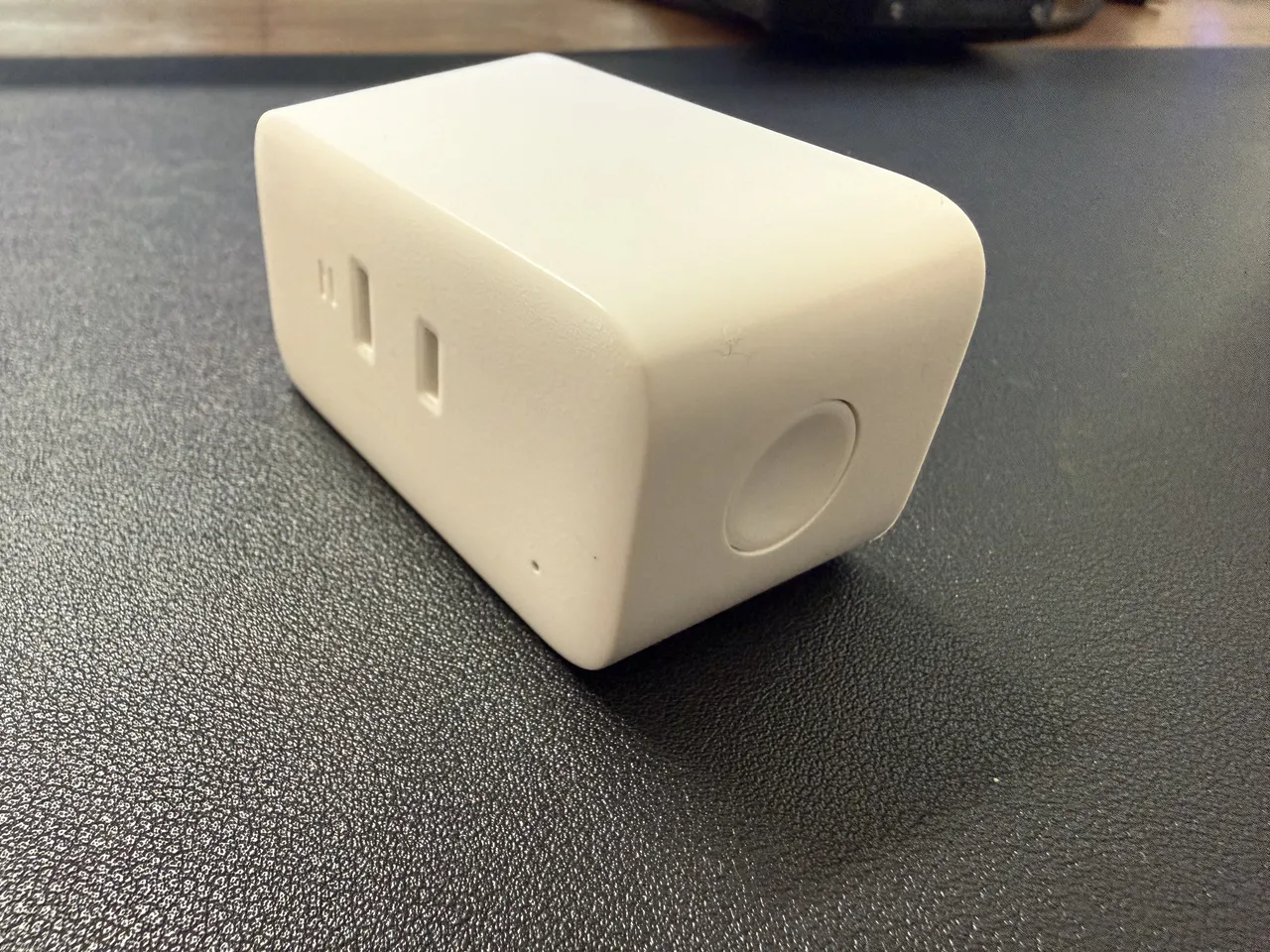 黒いデスクマットの上に置いた白い SwitchBot プラグミニを斜め上から。側面にコンセントの差し込み口、正面に丸いボタン
