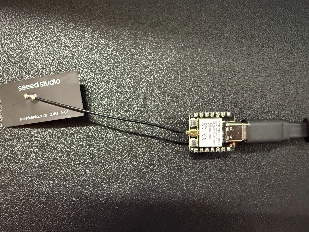 黒いデスクマットの上の XIAO ESP32C3。USB-C のケーブルがつながり、細いケーブルで外付けの板状のアンテナ(seeed studio 2.4G)につながっている
