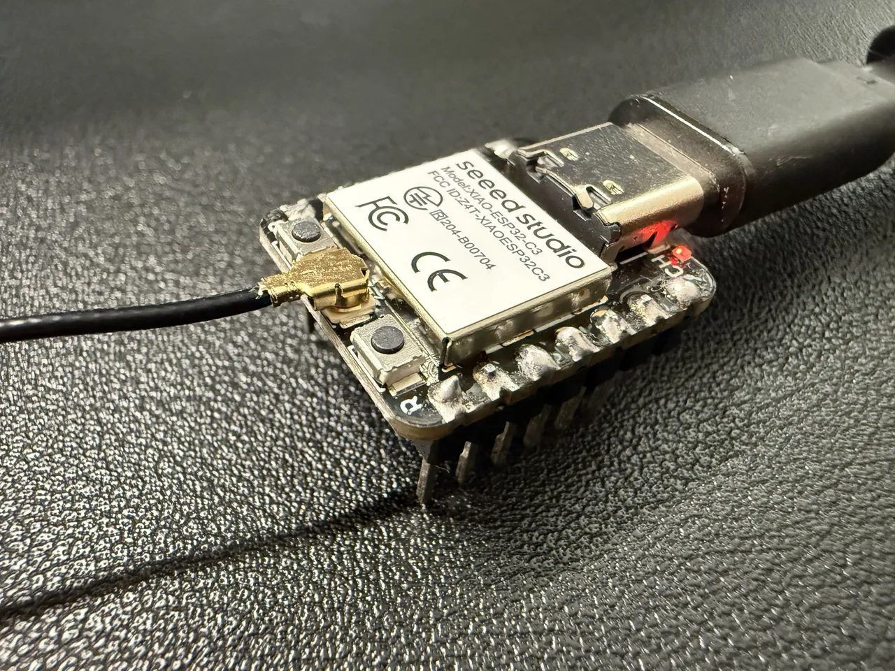 XIAO ESP32C3 の基板を斜めから寄って撮ったもの。金属のシールドに seeed studio と型番の表示があり、左の端子にアンテナのケーブルがつながり、右に USB-C のケーブルが挿さっている